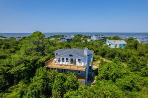 A coastal house with a deck, surrounded by dense greenery and a view of the sea. - Sandy Sea View: Soundside in Southern Shores with Ocean Views (Southern Shores)