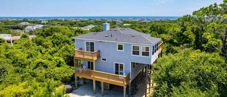 A two-story house with a balcony, surrounded by dense greenery and a clear view of the ocean.