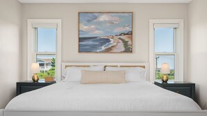 A bedroom with a large bed, two bedside tables with lamps, a painting of a beach scene, and windows with a view of greenery.