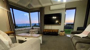A living room with a sofa, a coffee table, a flat-screen TV, and large windows offering a view of the outdoors.