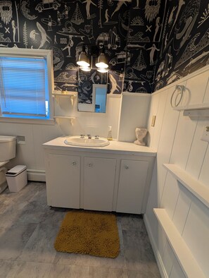 Un baño con tocador blanco, un lavamanos y un espejo. Las paredes están decoradas con un mural en blanco y negro. Hay una ventana con persianas y un perchero para toallas.