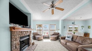 A living room with a stone fireplace, two brown leather sofas, a ceiling fan, and a large TV mounted on the wall. - Carolina Breeze - Your Perfect Getaway! (Merritt)