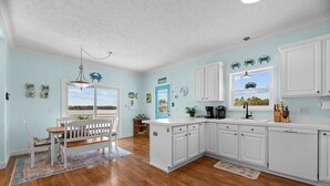 A kitchen with white cabinets, a wooden dining table with chairs, and a view of the outdoors through a window. - Carolina Breeze - Your Perfect Getaway! (Merritt)