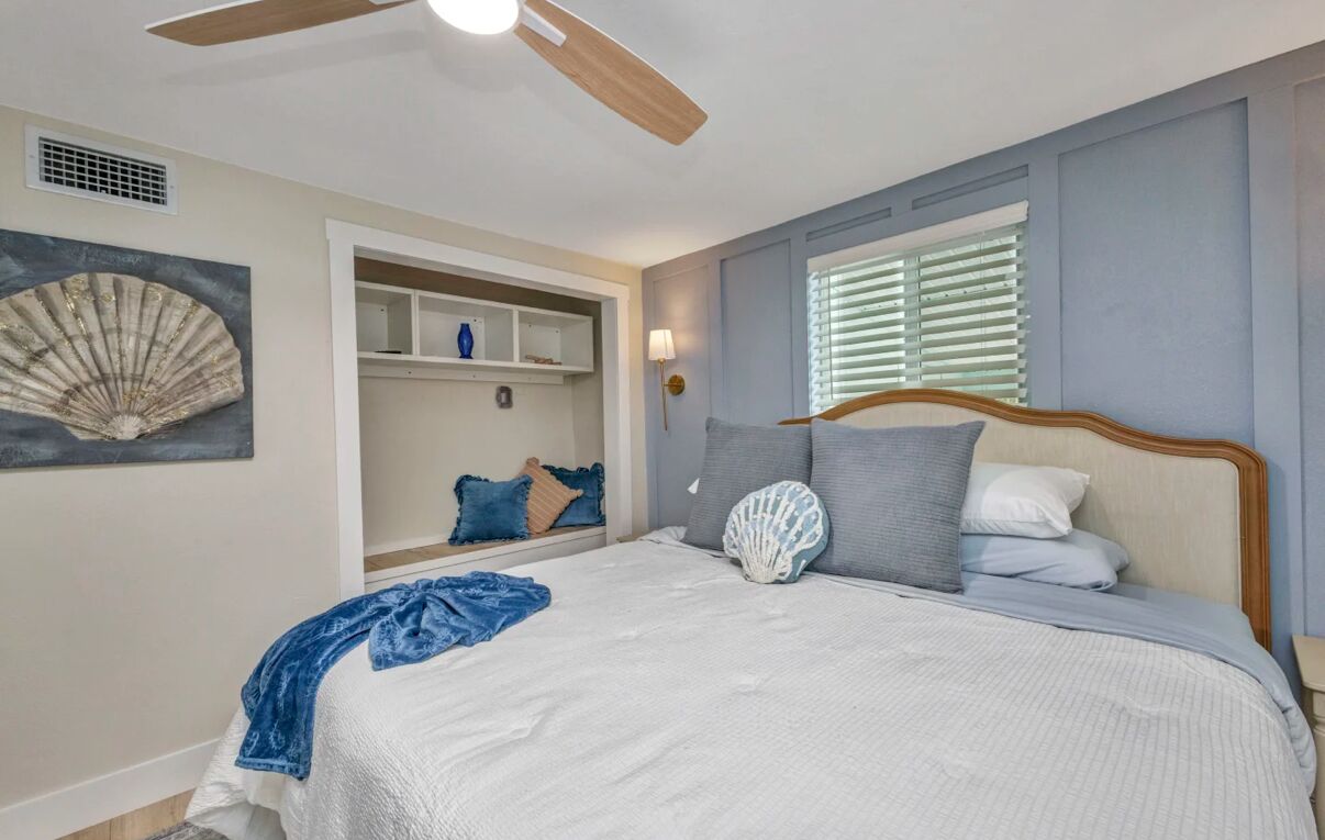 A bedroom with a bed, a ceiling fan, a window with blinds, a built-in shelf, and a decorative shell on the wall.