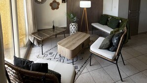 A modern living room with a sofa, a coffee table, a side table, and a wicker chair. There is a window with curtains, a wall-mounted mirror, and a decorative wall panel. - 2-Bedroom holidays Cottage with 2 Terrace, Air Conditioning  et  Parking (CANET PLAGE)