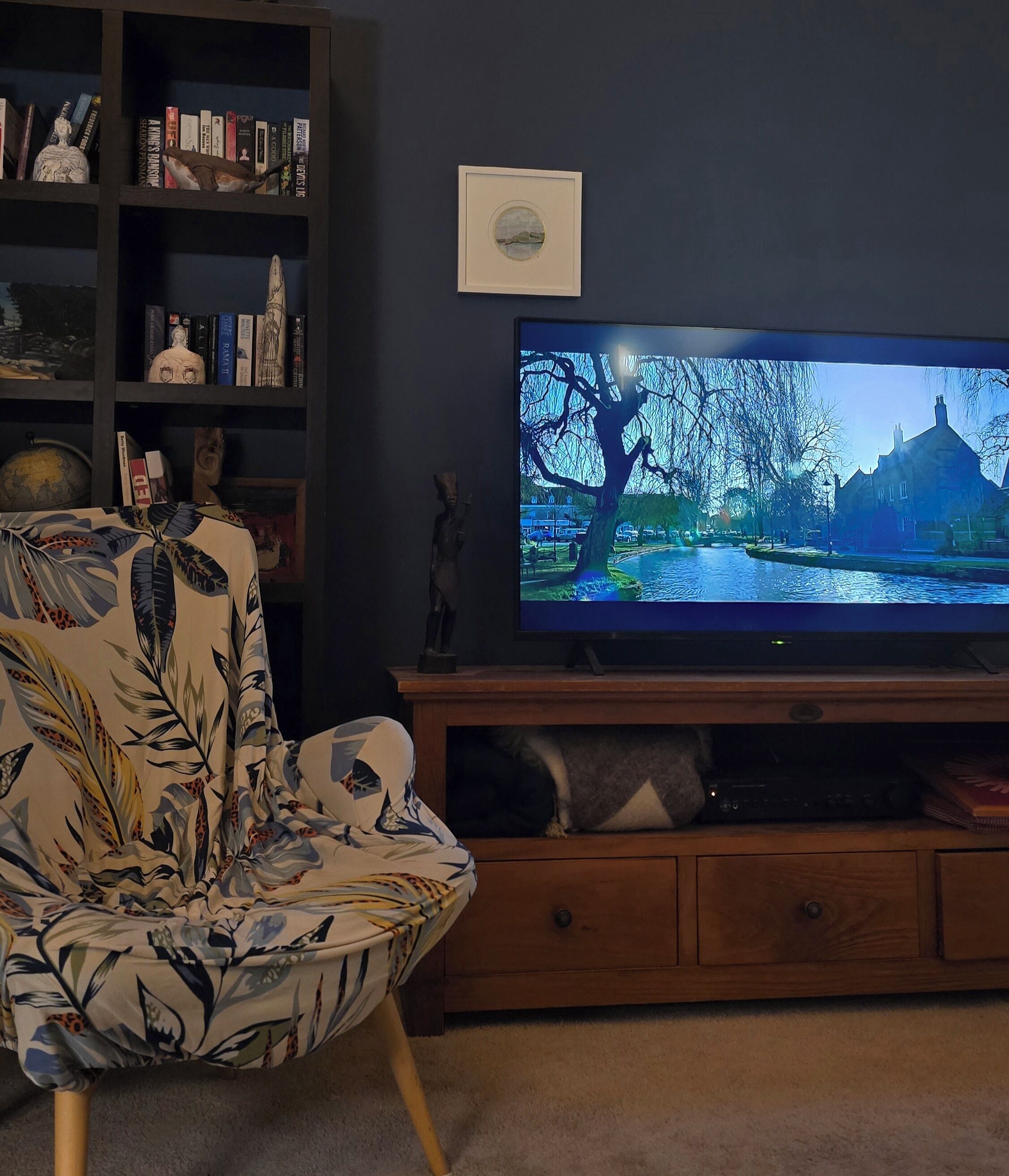 Un salon doté d’un grand téléviseur à écran plat, d’un meuble de rangement en bois, d’un fauteuil à motifs floraux et d’une bibliothèque remplie de livres et d’objets décoratifs.