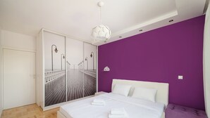 Een slaapkamer met een bed, een wit hoofdbord, paarse muren en een grote spiegel met uitzicht op een houten terras.