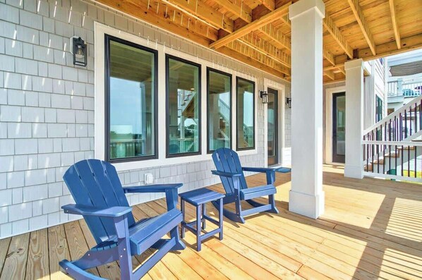 A covered outdoor area with wooden flooring, large windows, and blue Adirondack chairs. - Sweet Breeze by Brunswick Vacation Rentals: New 5BR w/ Elevator & Community Pool (Ocean Isle Beach)