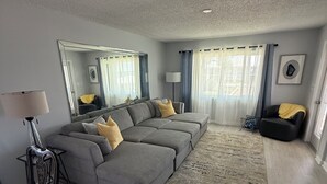 A modern living room with a grey sectional sofa, a glass coffee table, a floor lamp, and a large mirror. - Sunsets and Palm Trees overlooking the St. Lucie River (Stuart)