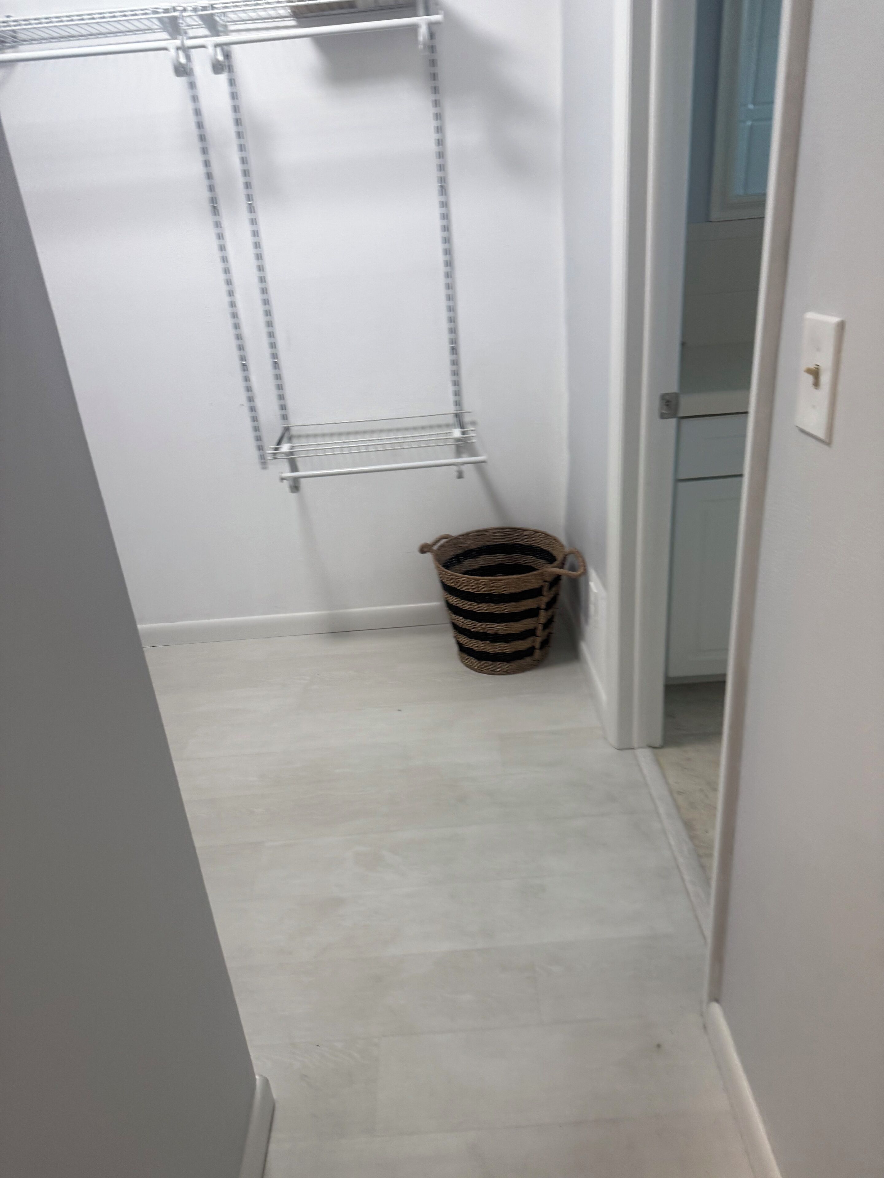 A narrow hallway with a white floor, a metal shelf unit on the left, and a wicker basket on the right.