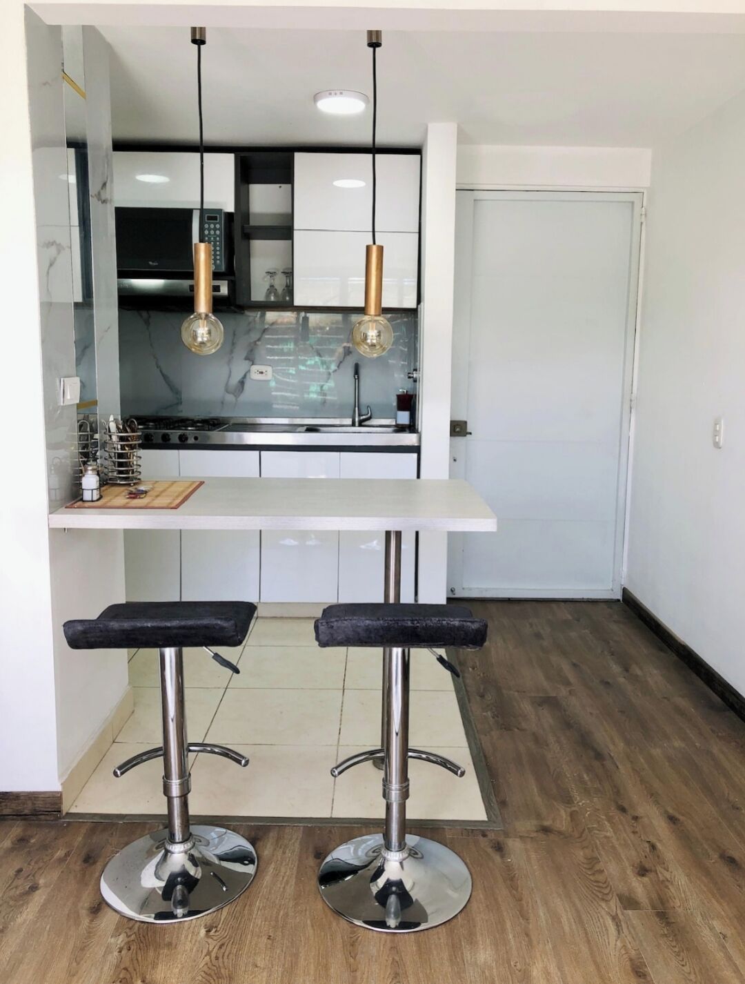 Une cuisine moderne avec un plan de travail blanc, deux tabourets de bar et des luminaires suspendus.