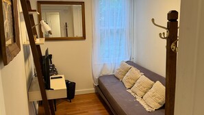 Habitació compacta amb terra de fusta, un sofà gris amb coixins estampats, un escriptori amb ordinador i làmpada, un mirall i una finestra amb cortines de tela fina.