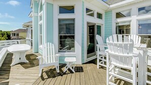 Um deck coberto com cadeiras Adirondack brancas, uma mesa pequena e uma porta de vidro que dá para um edifício com paredes externas azuis e brancas.