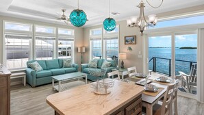 Un salón amplio con un ventanal grande con vista al mar, un conjunto de sofá color turquesa, una mesa de centro de madera y un comedor con vistas.