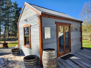 A rustic cabin with a wooden door, a window, and a small porch area with two barrels.