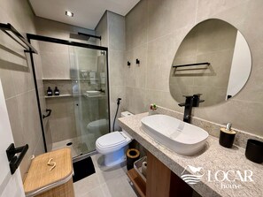 A modern bathroom with a glass-enclosed shower, a white toilet, a large sink with a mirror, and a small wooden stool.