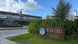 Bordje met 'Beach Class Summer Residence', inclusief logo, met op de achtergrond een helderblauwe lucht, een verhard pad en een modern gebouw.