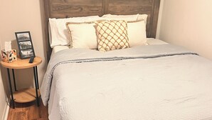 Una cama bien tendida con un edredón gris, un cabecero de madera y una mesita de noche con un marco de fotos y una caja de pañuelos.