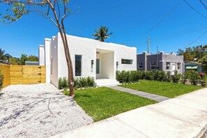 A modern two-story house with a white exterior, a front door, and a small front yard with a tree and shrubs. - Elegant designer retreat in Miami Design District with luxe vibes (Miami)