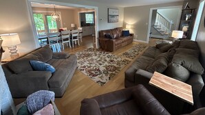 A spacious living room with a brown leather sofa set, a grey sofa, and a wooden coffee table. There is a dining area with a white table and chairs, and a kitchen in the background.