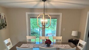 A dining area with a rectangular table, white chairs, a chandelier, and a window with a view of greenery.
