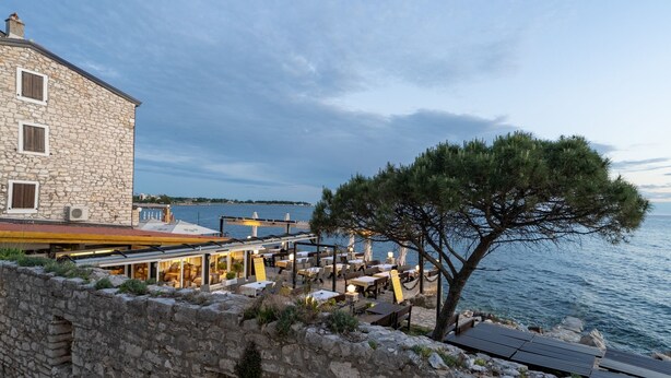 A stone building with outdoor seating by the sea, a tree, and a view of the water.