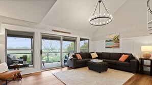 Ein geräumiges Wohnzimmer mit einer großen Eckcouch, einem Couchtisch und einem Kronleuchter. Große Fenster bieten einen Ausblick ins Freie.