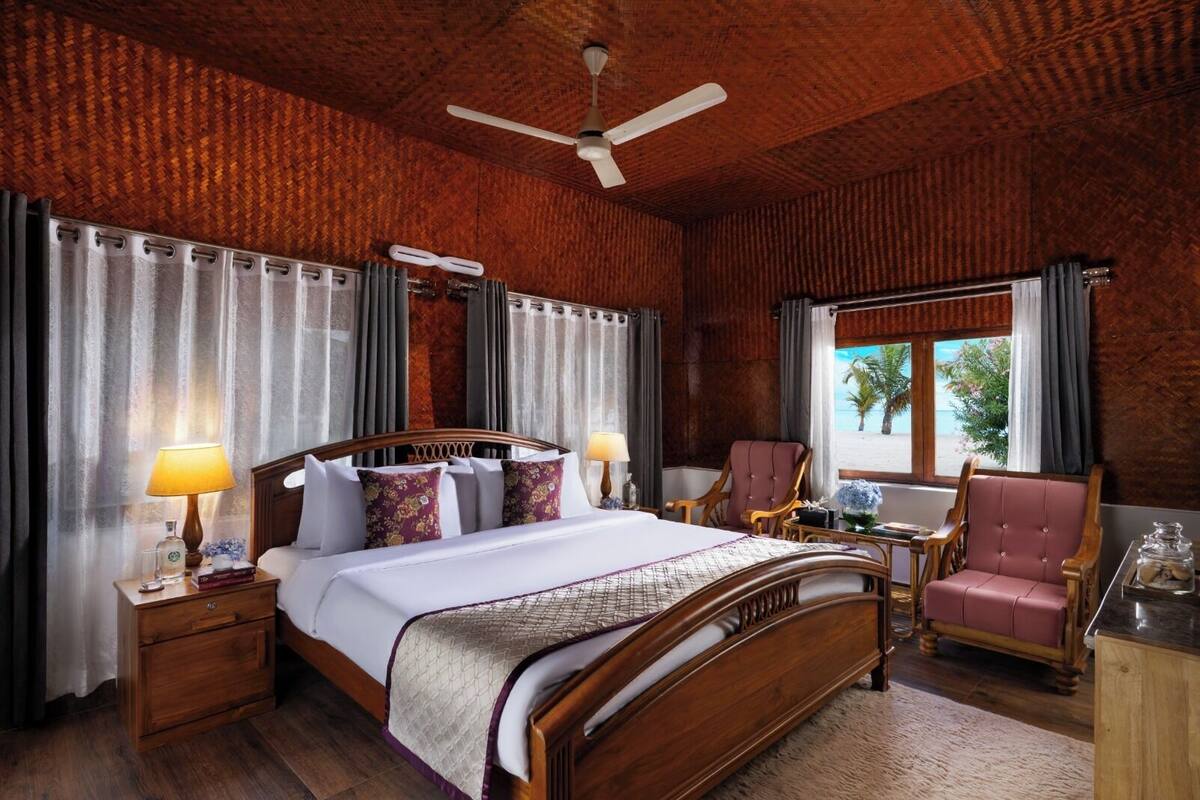 A spacious bedroom with a large bed, a wooden headboard, a ceiling fan, and a view of the beach through the window.