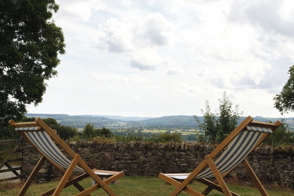 Deux fauteuils de jardin en bois, dotés de coussins à rayures, sont placés sur une pelouse, adossés à un mur de pierre, et offrent une vue sur un paysage pittoresque.