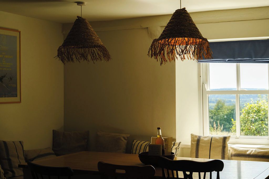A dining area with a round table and chairs, two hanging woven light fixtures, a window with a view, and a bottle on the table.