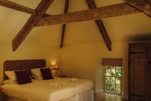 A bedroom with a bed, pillows, a nightstand with a lamp, a window with a view, and a wooden door.