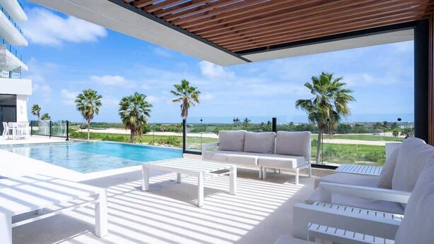 Un área de piscina moderna al aire libre, con una piscina blanca, sillas de descanso y vistas al océano y palmeras.