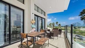 Un patio exterior moderno con mesa y sillas de madera, ventanas amplias y vistas a palmeras y un cielo despejado.