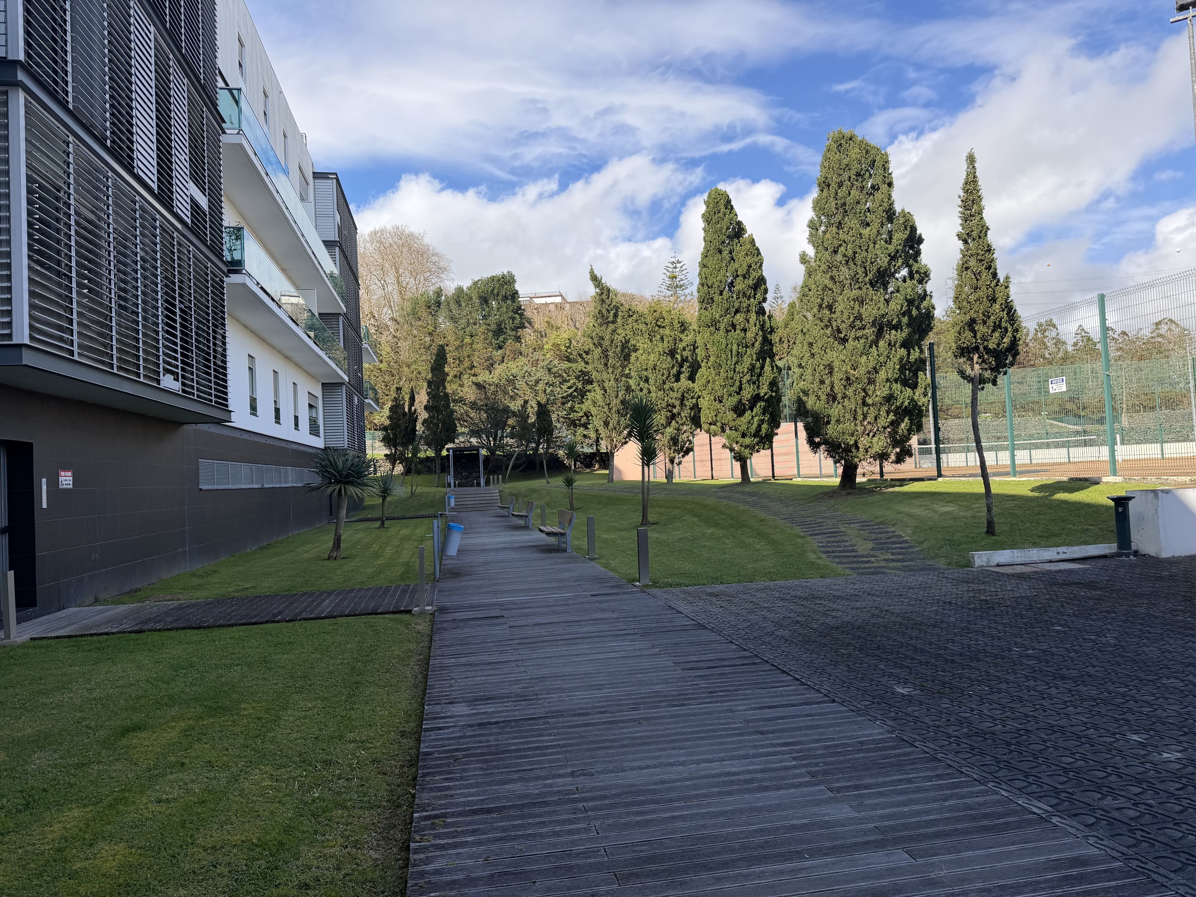 A pathway leading to a modern building with a flat roof, surrounded by a well-maintained lawn and tall trees.