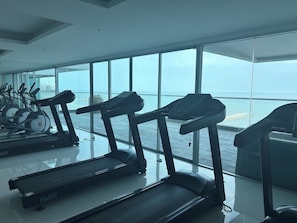 A gym with treadmills and exercise bikes, large windows, and a view of the sea.