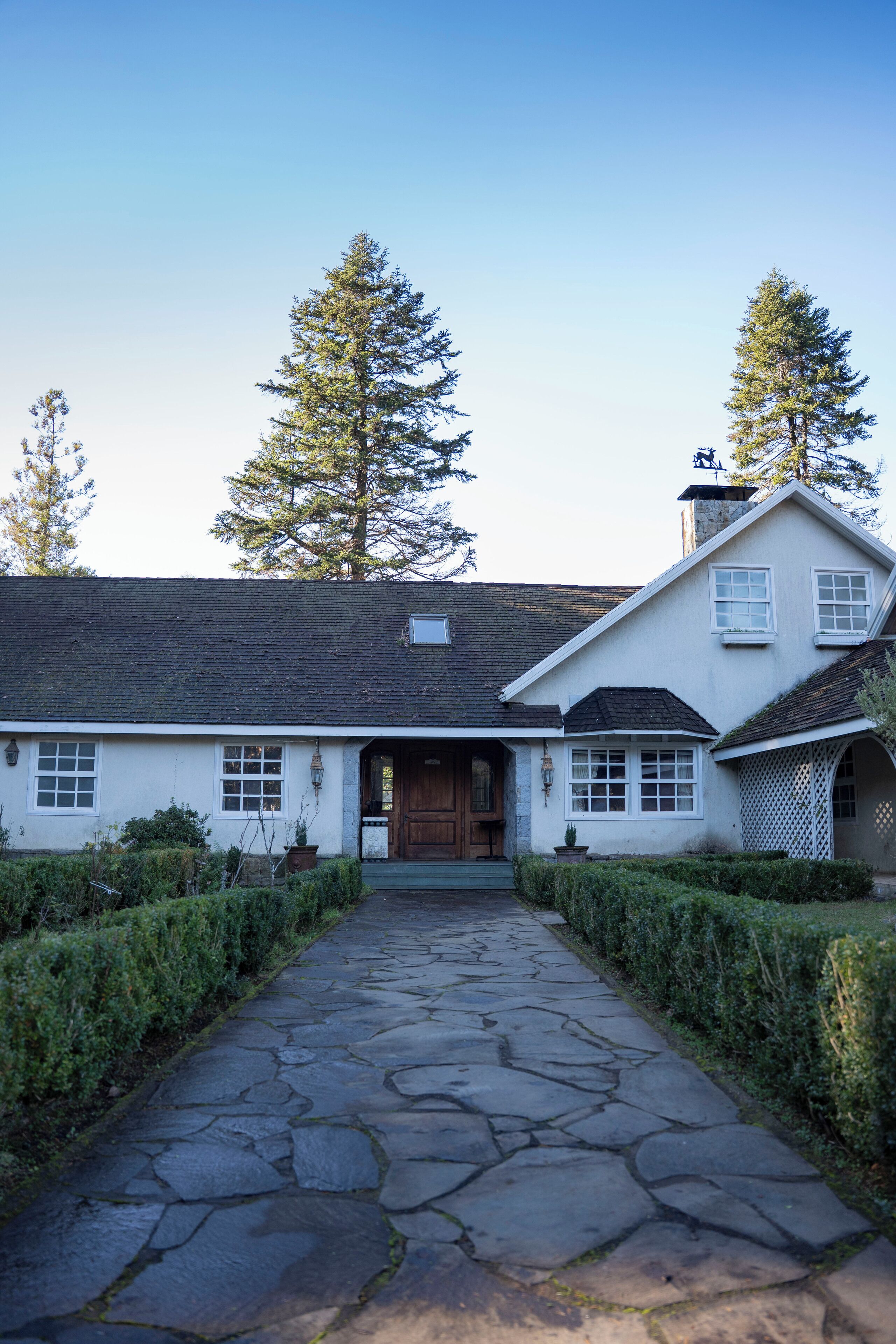 A traditional house with a stone pathway leading to the entrance, surrounded by neatly trimmed hedges and tall trees.