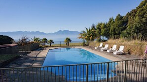 Une piscine rectangulaire entourée d’une clôture métallique, agrémentée de chaises longues et de palmiers, avec une chaîne de montagnes en arrière-plan.