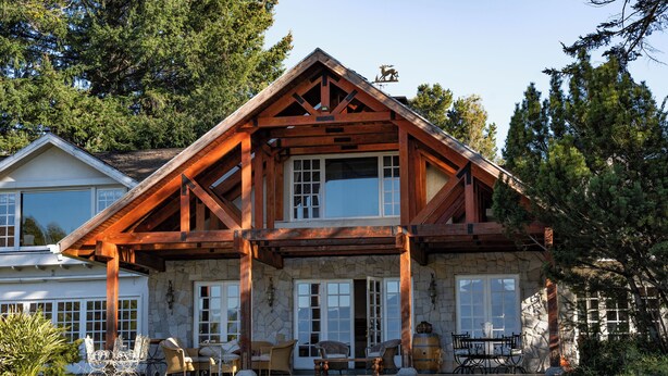 Une maison à deux étages avec une façade en pierre, des poutres en bois et une grande véranda à l’avant, aménagée avec du mobilier d’extérieur.