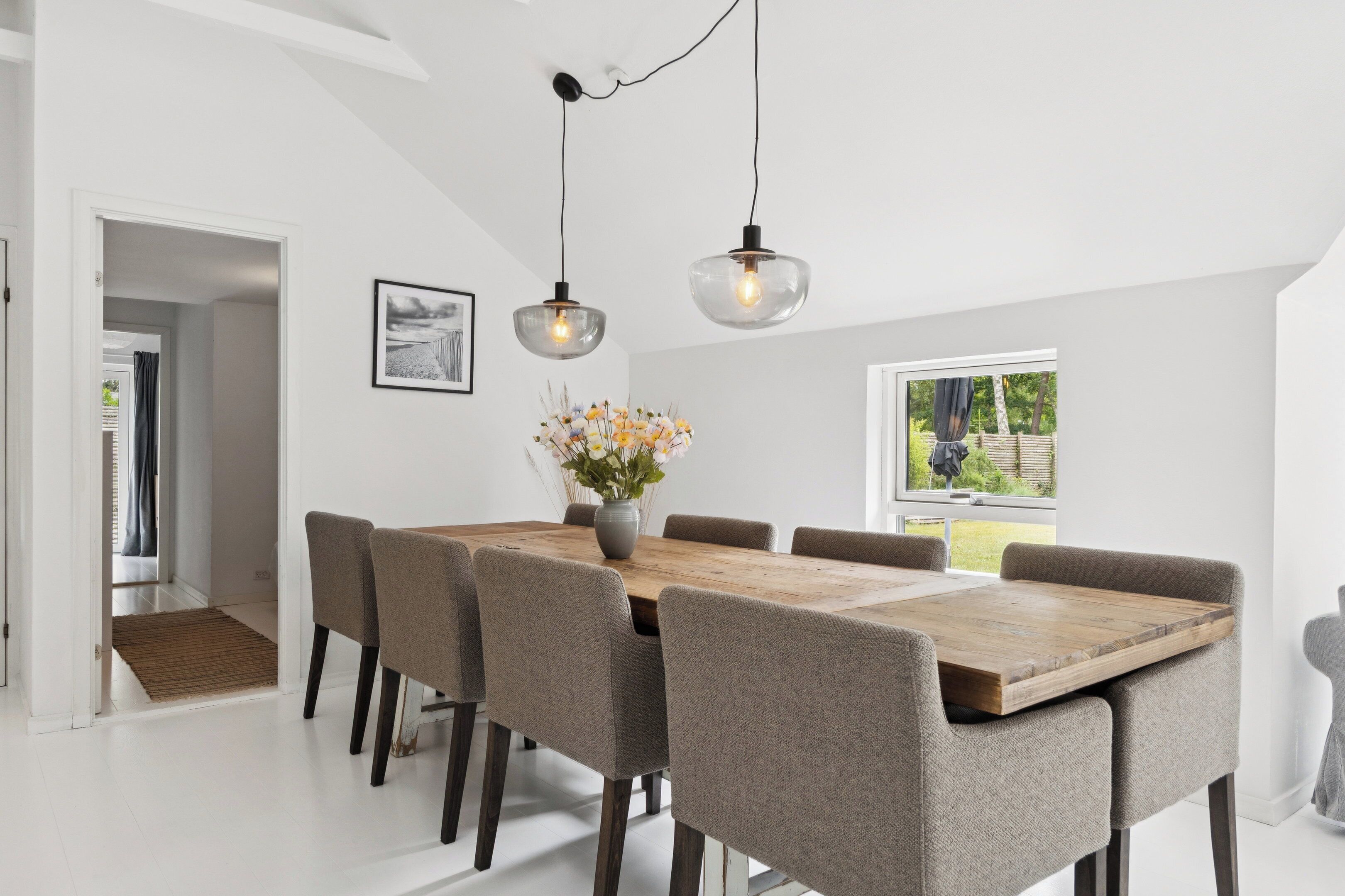 Et spiseområde med et træbord, seks stole, to hængende lamper, en vase med blomster og et vindue med udsigt til grøn bevoksning.