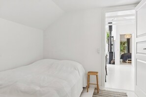 A minimalist bedroom with a bed, a small stool, and a doorway leading to another room. - Newly renovated holiday home near the beach (Væggerløse)