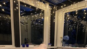 A cozy room with a wooden floor, a white chair, a small table, a lamp, and a window adorned with string lights. - A cozy older summerhouse on a small island (Orø)