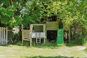 Una casetta gioco in legno con una scala e una porta verde, circondata da alberi ed erba.