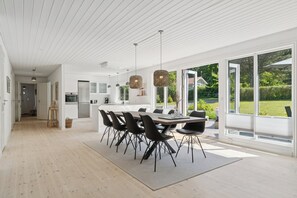 A modern kitchen with white cabinets, a dining area with a large table and chairs, and large windows offering a view of the outdoors.