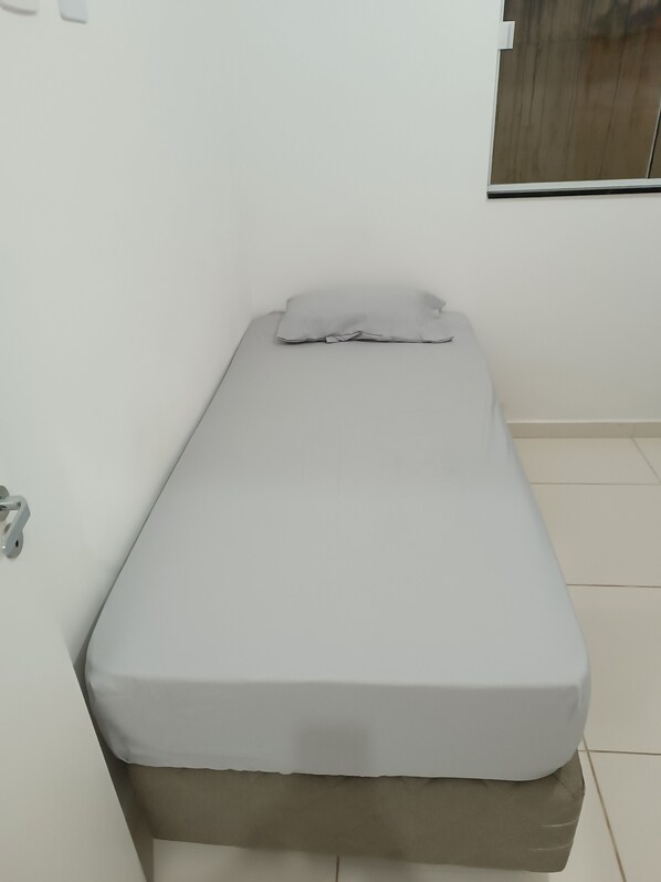 A single bed with a grey mattress and pillow in a room with white walls and a window.