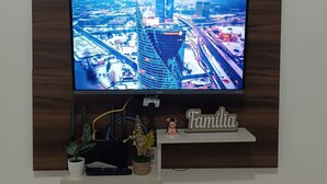 A flat-screen TV mounted on a wooden wall, displaying a cityscape. Below the TV, there is a shelf with a small plant, a decorative item, and a sign that reads "Familia". - House in Matinhos with free Wi-Fi and accepts your pet. (Matinhos)