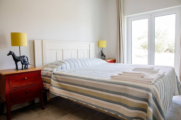 A neatly made bed with striped bedding, a wooden nightstand with a red drawer, two bedside lamps, and a window with curtains. - CASA DA BEIRÃ T2, country house (Marvão)