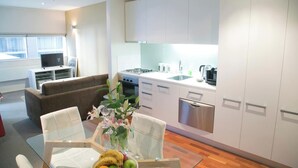 Een moderne keuken met witte kasten, een roestvrijstalen vaatwasser en een koffiezetapparaat. Er staat een glazen eettafel met een bloevas en een schaal fruit.