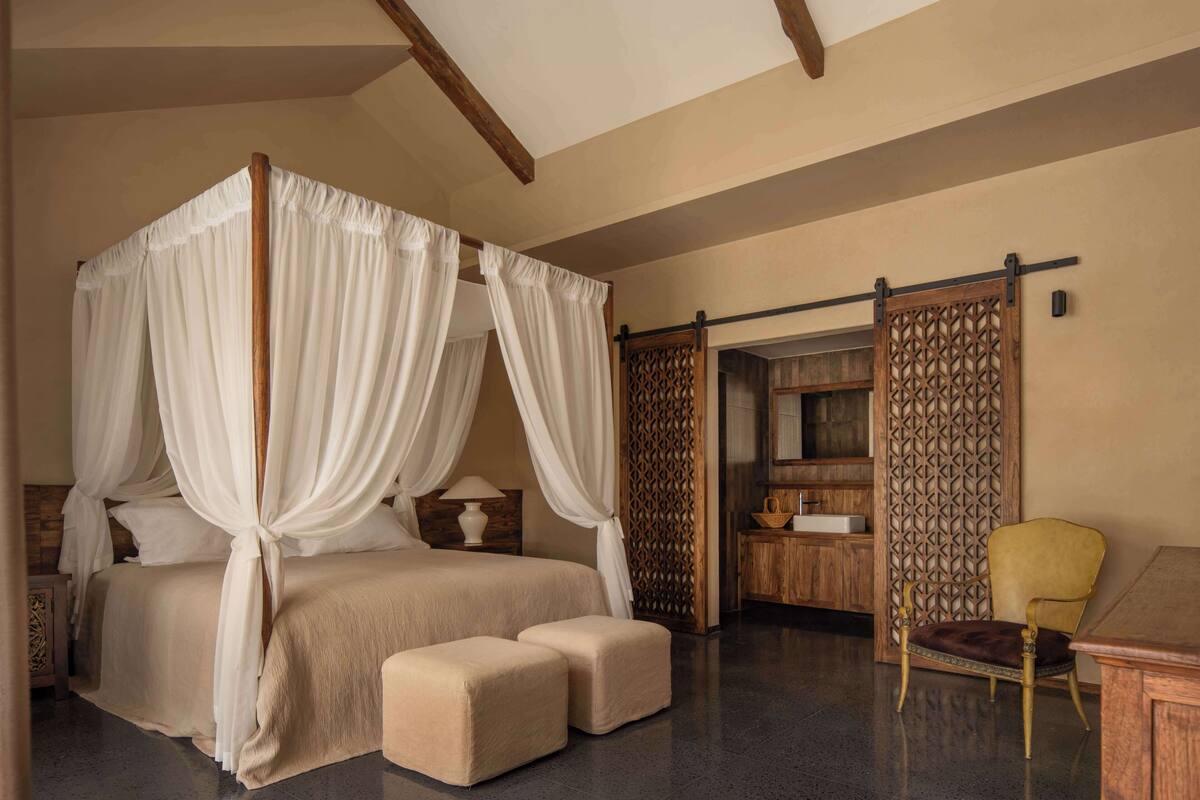A four-poster bed with white drapes, a wooden nightstand, a bedside table with a lamp, and a chair in a modern room with a wooden partition and a small table.