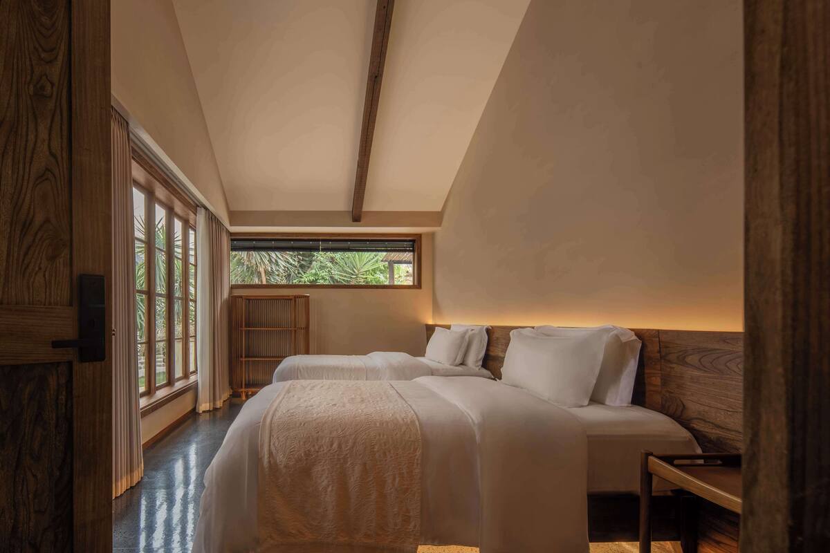 A bedroom with two beds, wooden headboards, a large window with curtains, and a view of greenery outside.