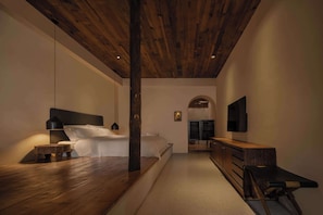 Une chambre avec un plafond en bois, un lit recouvert de draps blancs, une table de chevet avec une lampe et une commode avec un miroir.